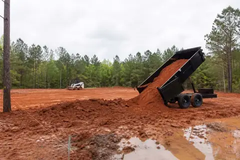 Dump trailer delivering red sandy fill while a skid steer spreads the material across a marked East Texas low spot